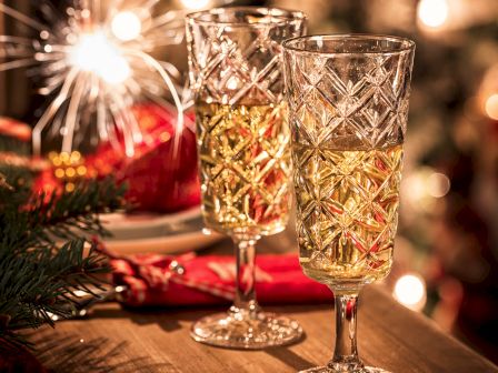 Two ornate glasses filled with a golden beverage sit on a table, with a festive, blurred background of lights and decorations.