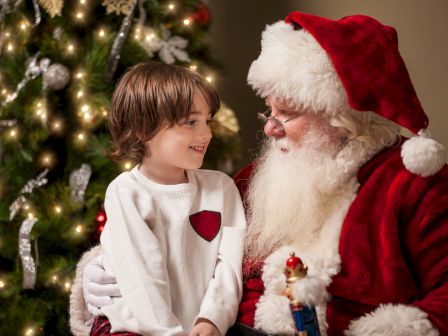 A child sits on Santa's lap, smiling beside a Christmas tree decorated with lights and ornaments.