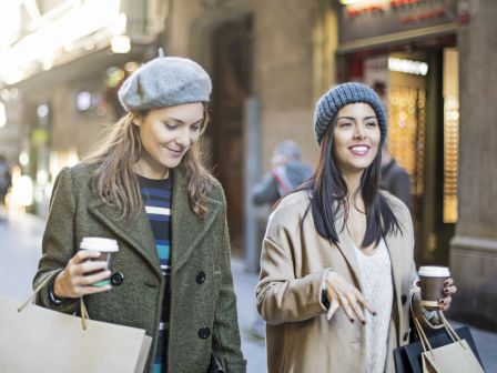 Two women walking on a city street, holding shopping bags and coffee cups, wearing coats and beanies, smiling and chatting.