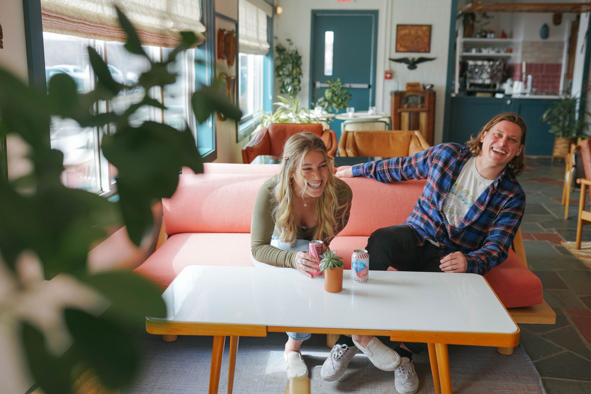 Two people sitting and laughing on a pink couch, with drinks on a table, in a cozy room with plants and a blue door.