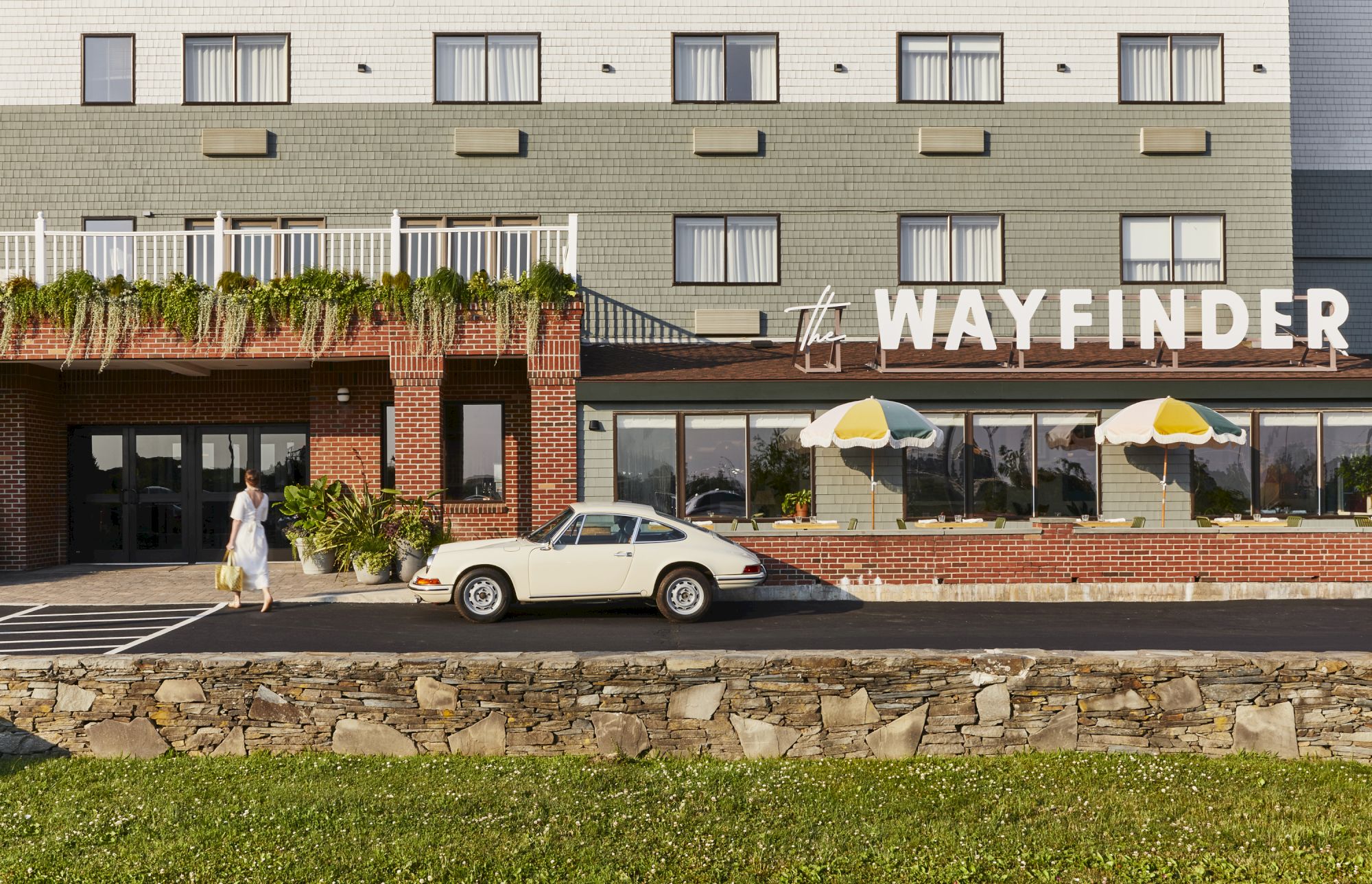 A person walks toward a beige vintage car parked in front of "The Wayfinder" hotel, with green and white walls and striped umbrellas.