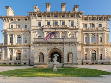 A grand, historical building with intricate architecture and an American flag displayed prominently at the entrance, surrounded by greenery.