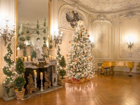 A lavishly decorated room with a large Christmas tree, ornate fireplace, candles, and a grand piano, all adorned for the holiday season.