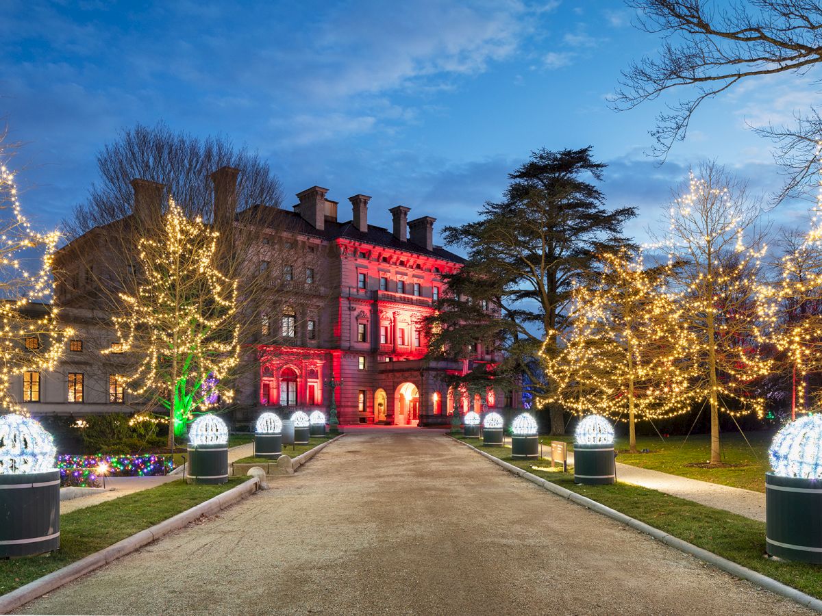 A grand building with festive lights illuminating trees and pathways, under a twilight sky.