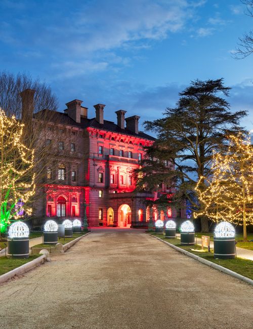 A grand building with festive lights illuminating trees and pathways, under a twilight sky.