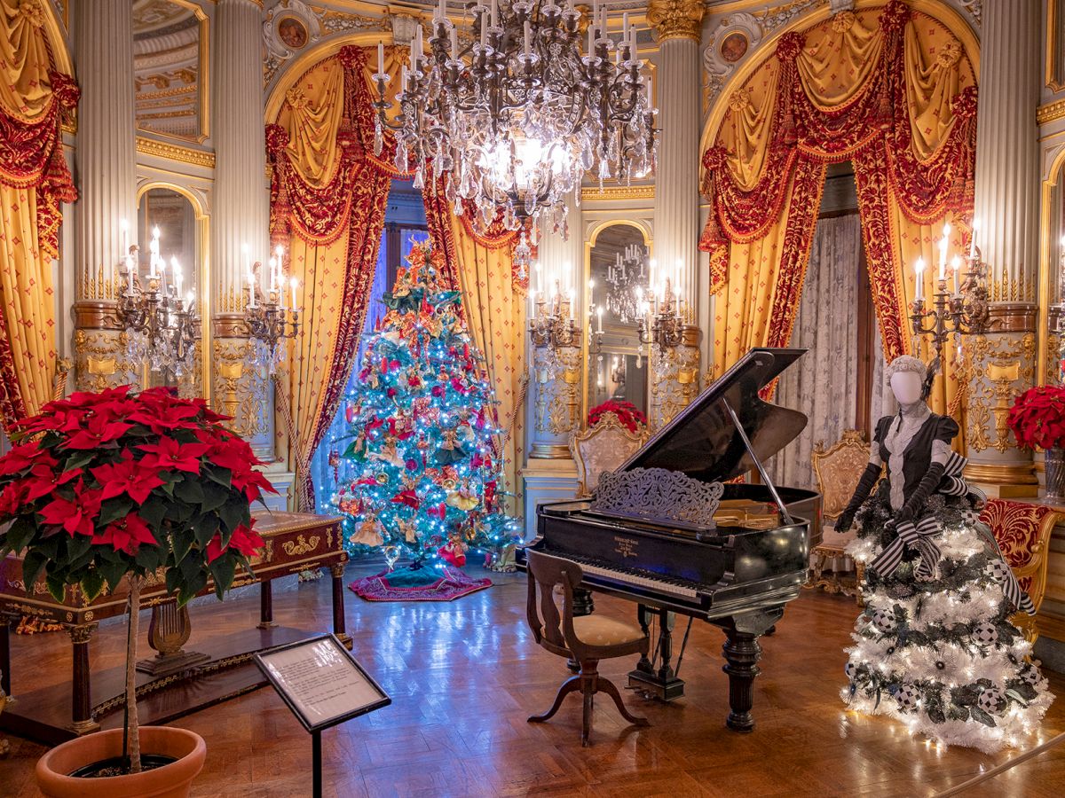 Ornate room with chandeliers, a decorated Christmas tree, a grand piano, mannequin, and red curtains. Festive and elegant setting.