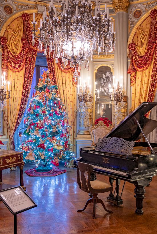 Ornate room with chandeliers, a decorated Christmas tree, a grand piano, mannequin, and red curtains. Festive and elegant setting.