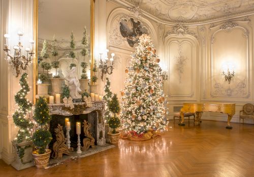 Elegantly decorated room with a lit Christmas tree, ornate fireplace, candles, greenery, and a grand piano in a luxurious setting.