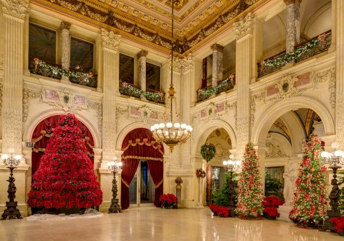 A grand hall decorated for Christmas with elaborate decorations, red ornamental trees, and classical architecture.