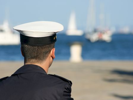 A person in a uniform with a white hat looks towards the sea, with blurred boats in the background.