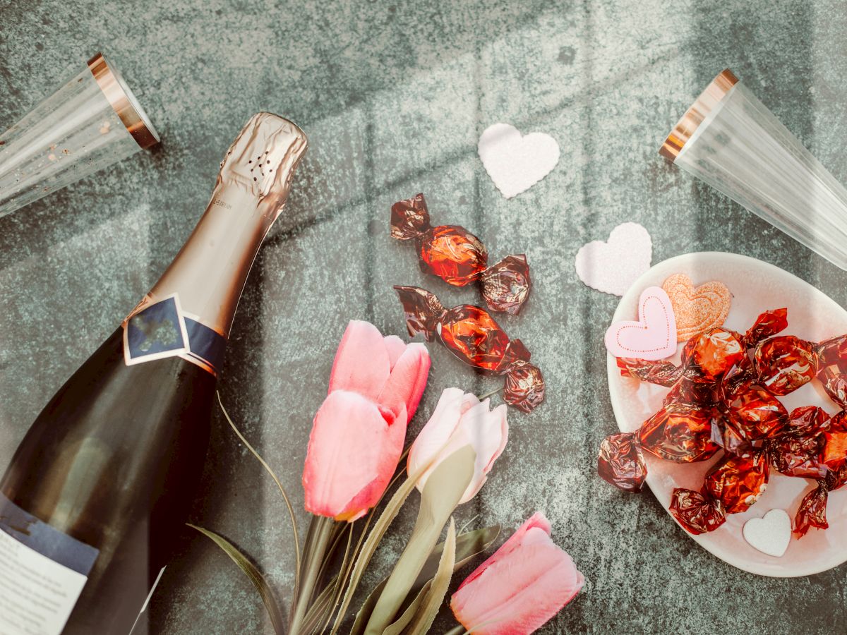 A bottle of champagne, two glasses, pink tulips, heart cutouts, and a plate of chocolates are arranged on a surface.