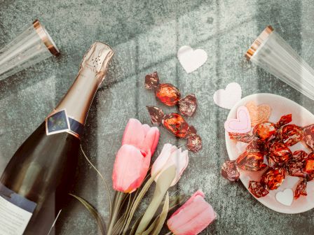 A bottle of champagne, two glasses, pink tulips, a plate of wrapped chocolates, and heart-shaped cutouts are on a textured surface.