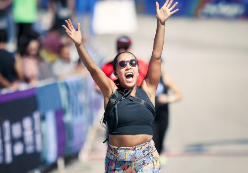 A person is joyfully crossing the finish line of a race, arms raised, wearing sunglasses and colorful athletic wear.