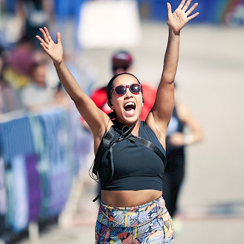 A person is joyfully crossing the finish line of a race, arms raised, wearing sunglasses and colorful athletic wear.