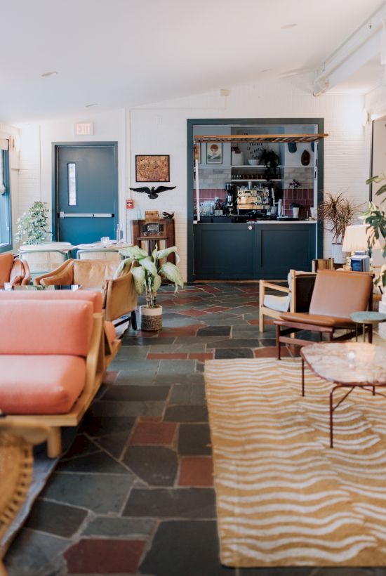 The image shows a stylish lounge with eclectic furniture, a coffee bar in the back, and warm lighting, creating a cozy atmosphere.