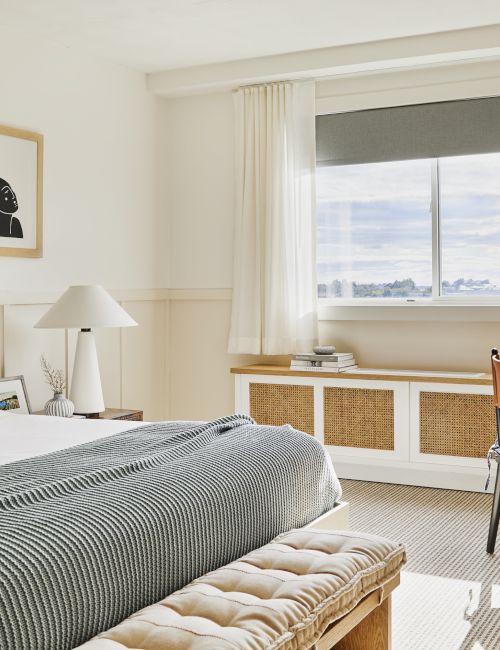 A cozy bedroom with a bed, chair, and table. Large window offers a view. Decor includes art, a blanket, and a woven lamp.