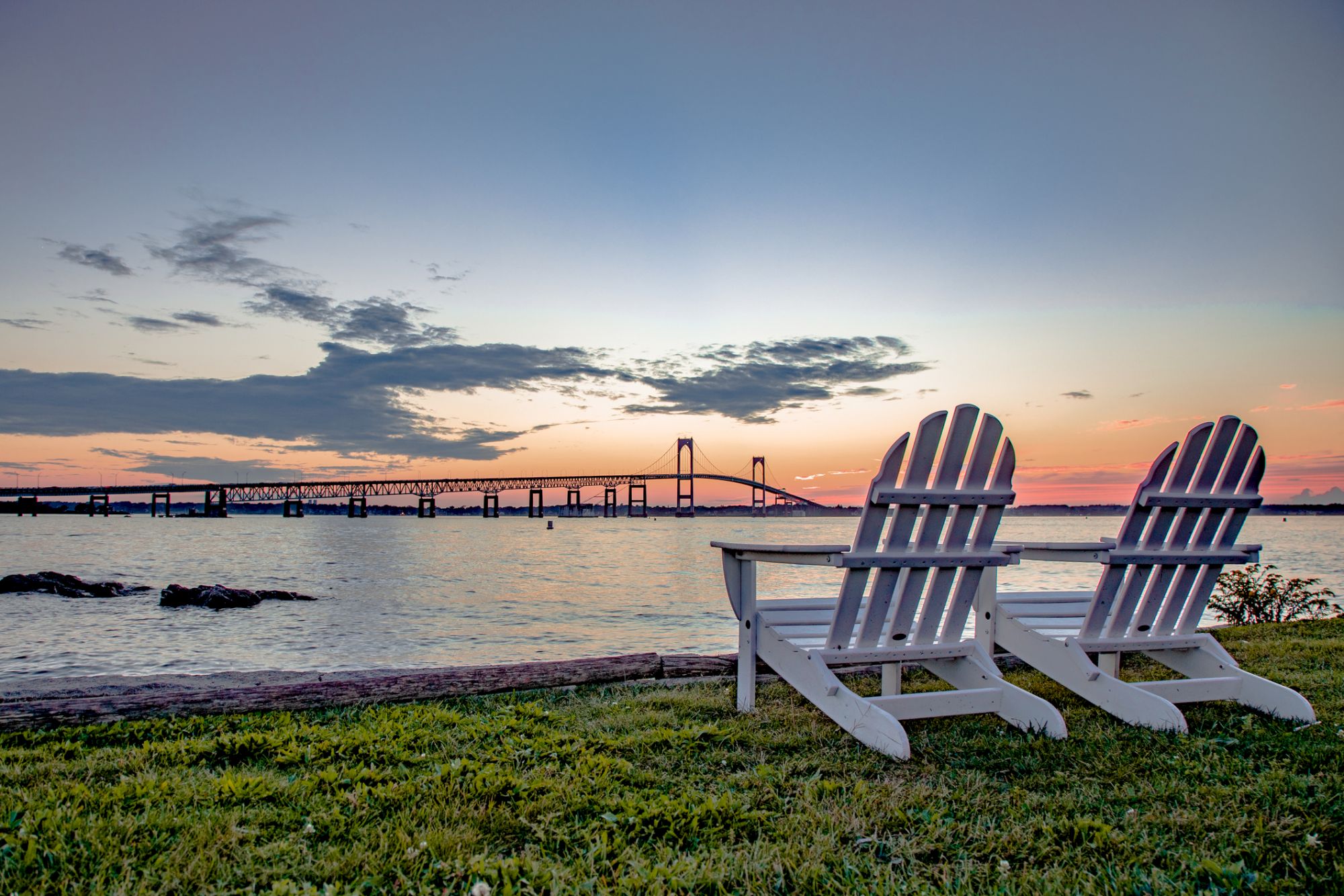 Two white Adirondack chairs face a scenic view of a bridge over calm water at sunset, with a tranquil sky and grassy foreground.