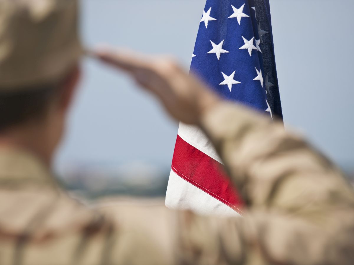 A person in military uniform saluting the American flag, with a blurred background.