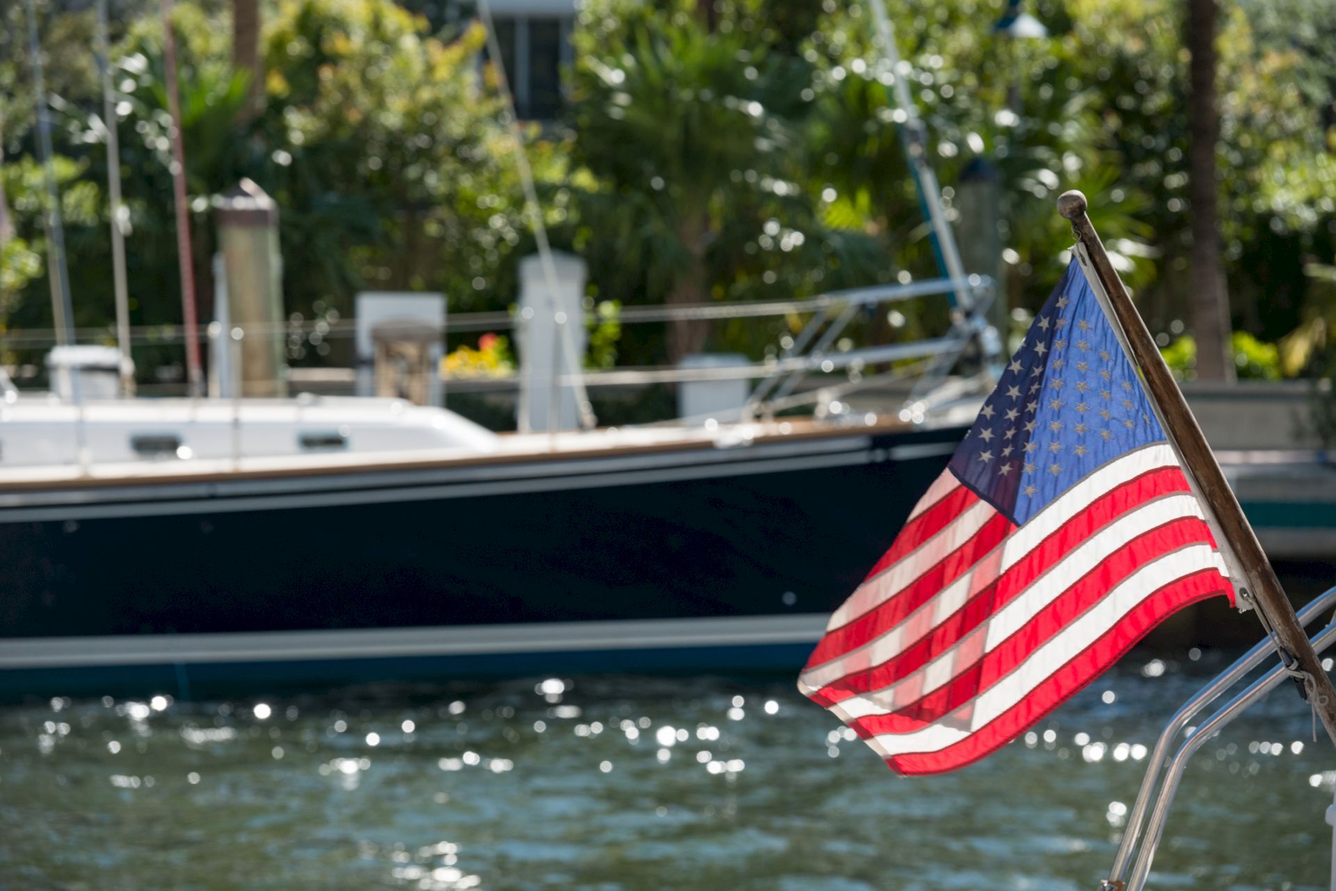 A US flag on a boat waves in the foreground with another boat and greenery visible in the background by the water.