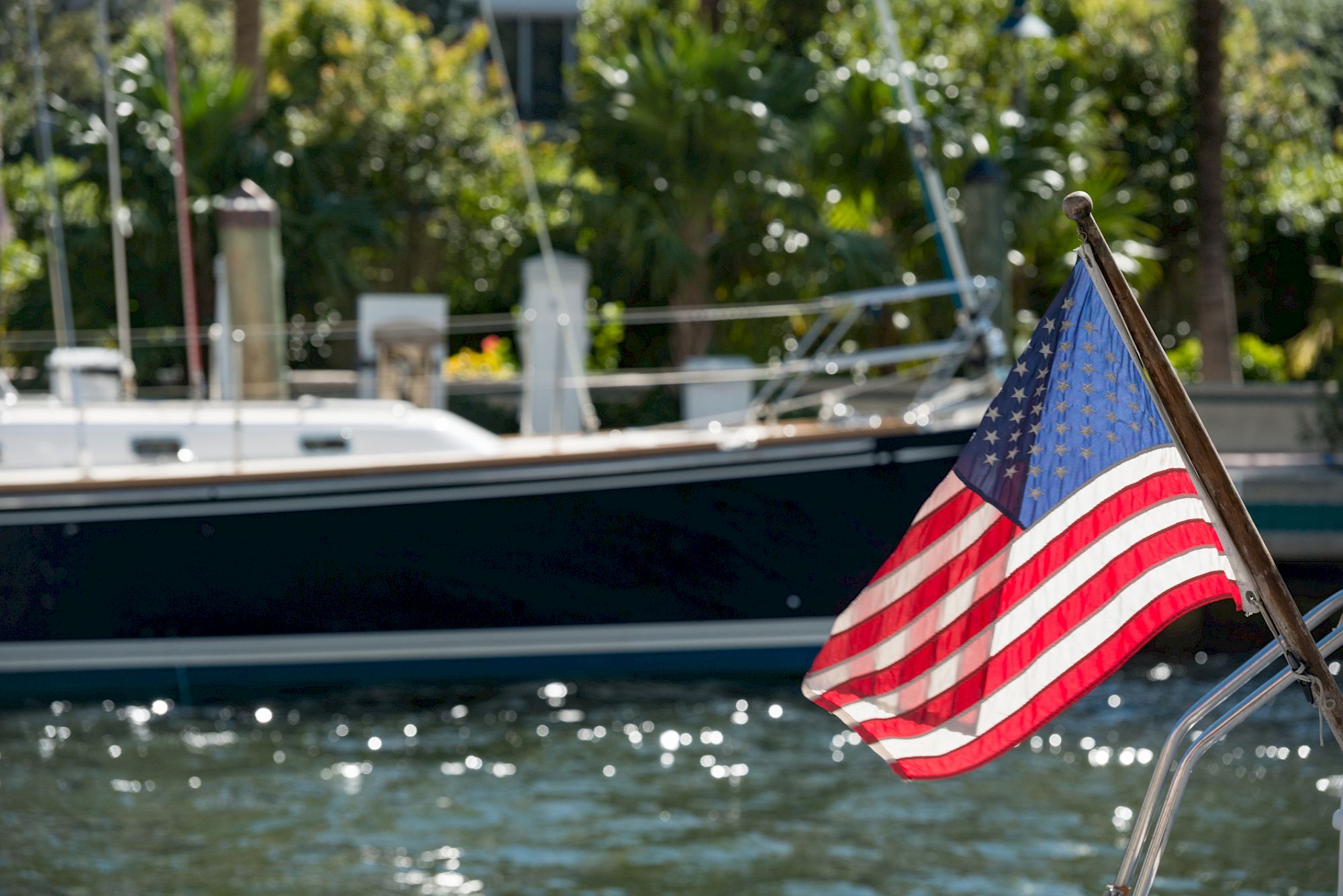 A US flag on a boat waves in the foreground with another boat and greenery visible in the background by the water.
