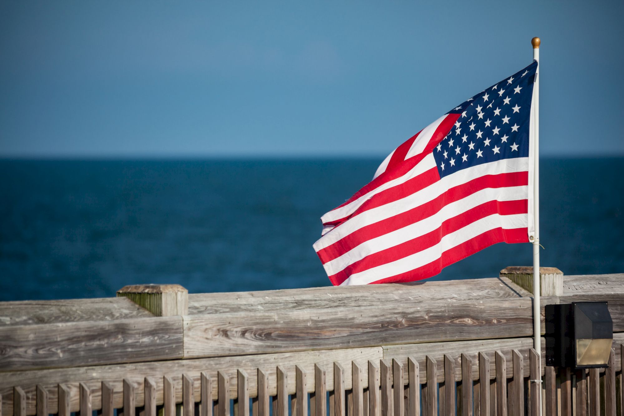 A U.S. flag waves on a pole by a wooden fence with the ocean in the background under a clear blue sky.