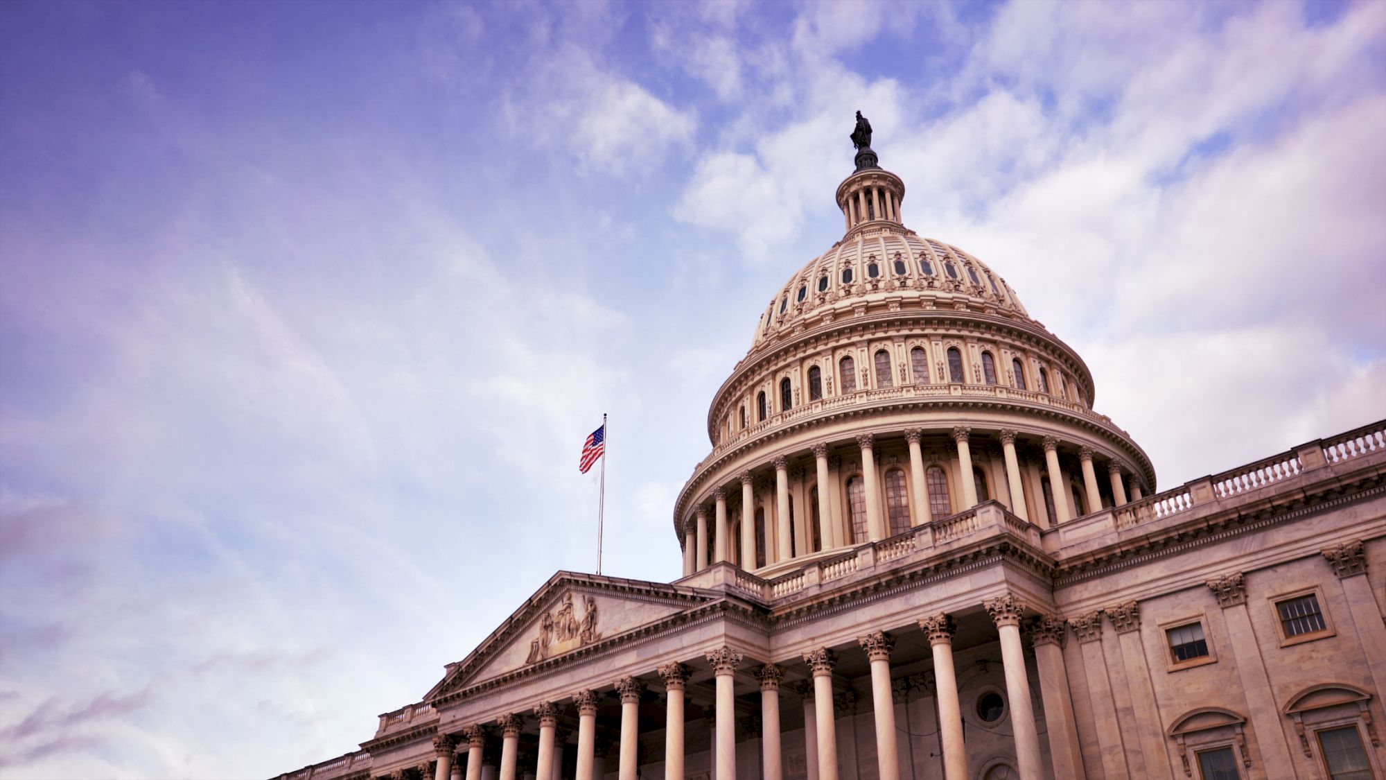 The image shows the United States Capitol building with its iconic dome, columns, and a waving American flag under a partly cloudy sky.