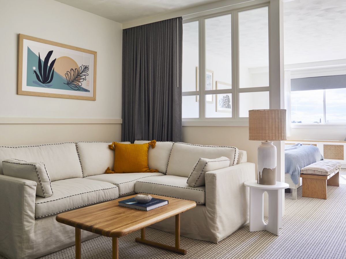 A cozy living area with a beige sectional sofa, a wooden coffee table, and a lamp. Decorative art and a window in the background complete it.