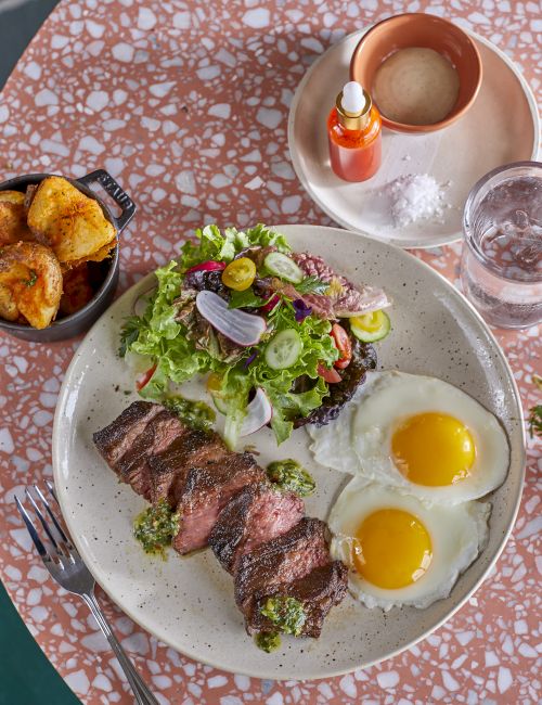 A plate with steak, salad, and sunny-side-up eggs, alongside roasted potatoes, a drink, hot sauce, and a glass of water on a terrazzo table.
