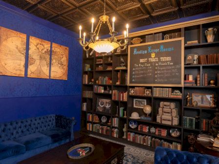 The image shows a cozy library with blue walls, tufted sofas, a chandelier, world map art, and bookshelves filled with books and decorative items.