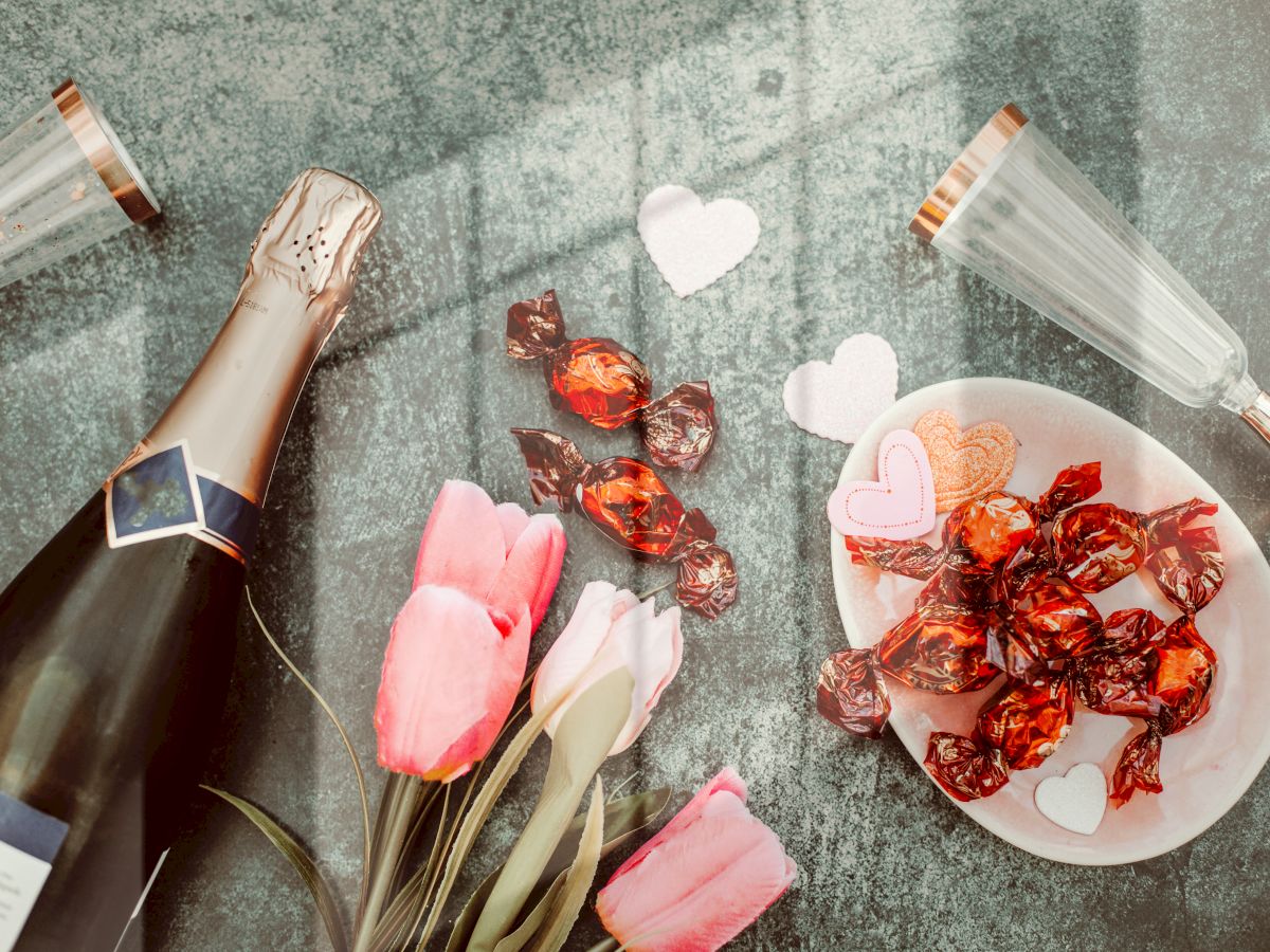 A bottle of champagne, two glasses, a bouquet of pink tulips, wrapped chocolates, and heart-shaped cutouts are arranged on a textured surface.