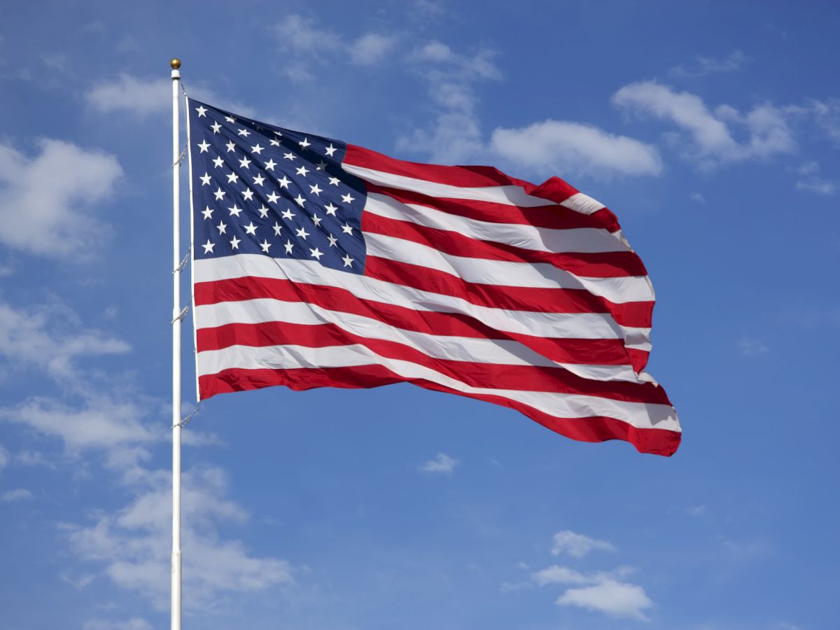 The image shows the United States flag waving against a blue sky with clouds.