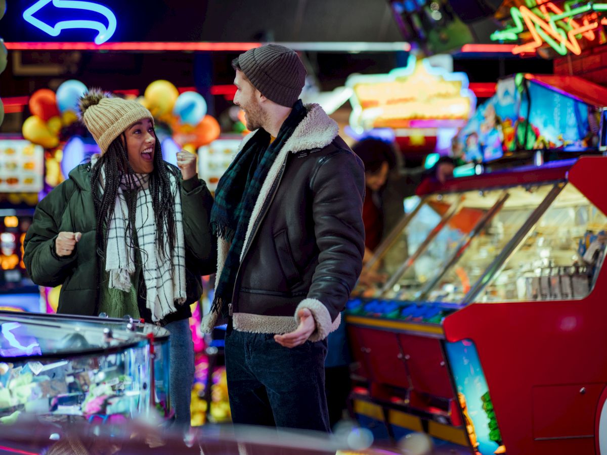 A man and woman in winter clothing are excitedly talking in a colorful arcade setting with various games and bright lights around.