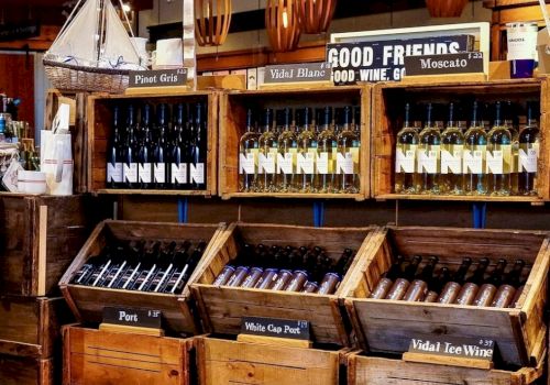 The image shows wooden crates displaying assorted wine bottles in a store with signs like "Pinot Gris" and "Vidal Blanc."