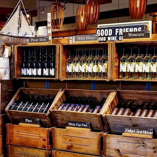 The image shows wooden crates displaying assorted wine bottles in a store with signs like "Pinot Gris" and "Vidal Blanc."