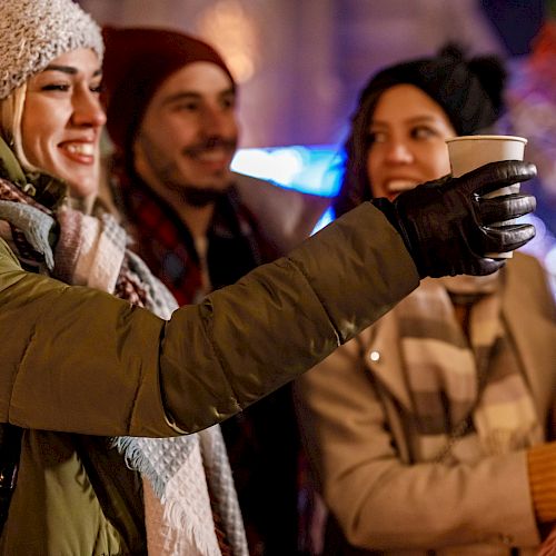 People bundled in winter clothes enjoying warm drinks at a festive outdoor event, with lights in the background.