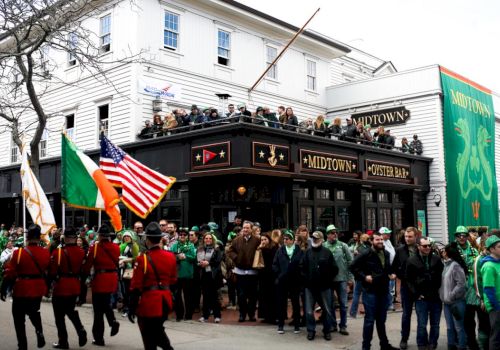 The image shows a parade with people dressed in green, flags, and a crowd outside Midtown Oyster Bar during a festive event.