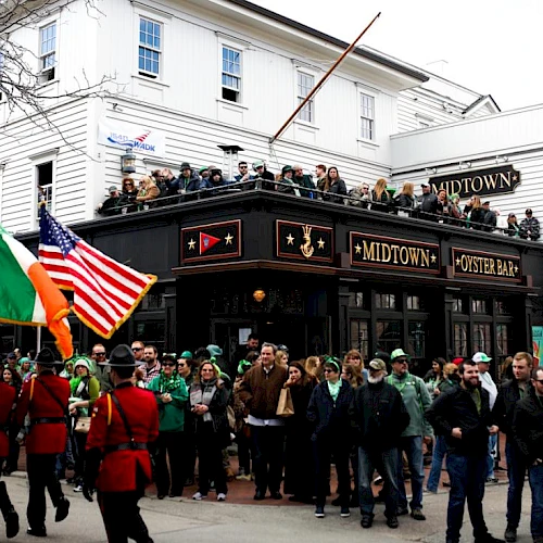 A street scene outside a pub with marching soldiers in red uniforms saluting as onlookers gather along storefronts and flags wave.