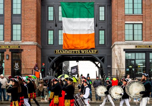 A lively parade in front of a brick building with a large Irish flag, featuring drummers and dancers.