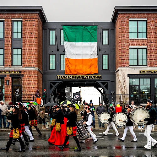 A lively parade in front of a brick building with a large Irish flag, featuring drummers and dancers.