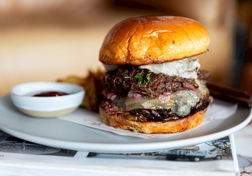 A juicy burger with melted cheese, onions, and greens on a glossy bun, served with a small side of sauce on a white plate.