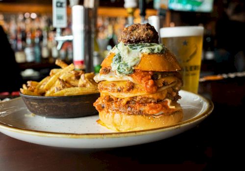 A stacked burger with various toppings, served with a side of fries on a plate. A pint of beer is in the background on a bar counter.