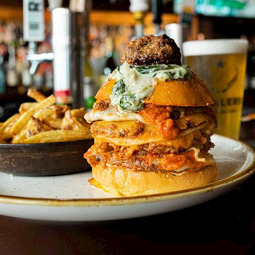 A stacked burger with various toppings, served with a side of fries on a plate. A pint of beer is in the background on a bar counter.