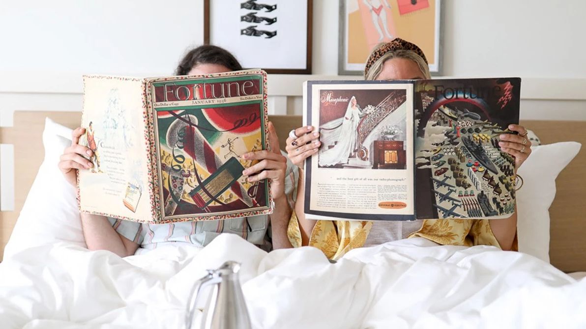 Two people are sitting in bed, each holding and reading a magazine.