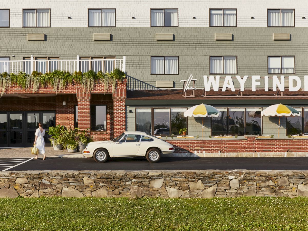 A woman walks near a vintage white car outside a building called "WAYFINDER," with umbrellas on a patio and greenery in front.
