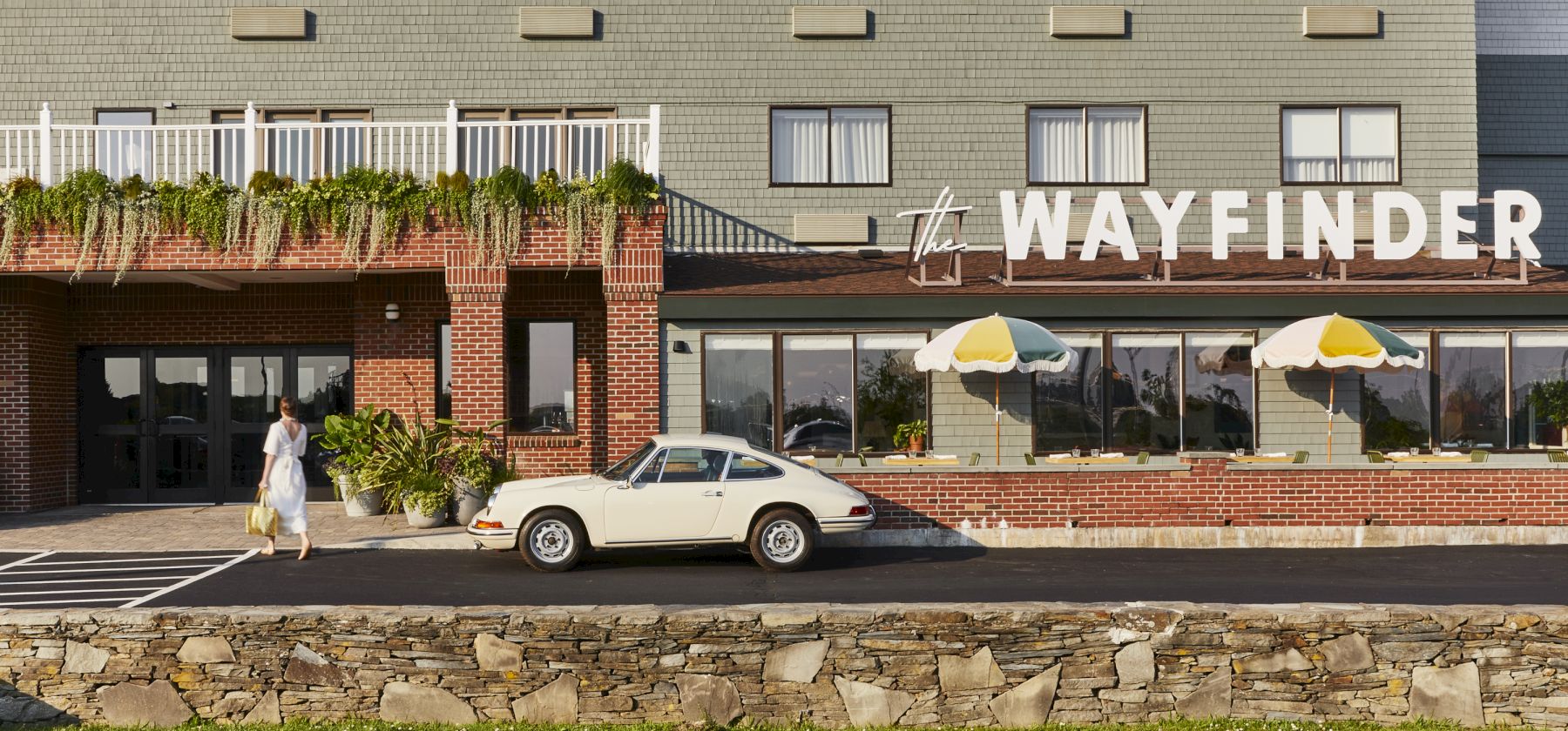 A woman walks near a vintage white car outside a building called "WAYFINDER," with umbrellas on a patio and greenery in front.