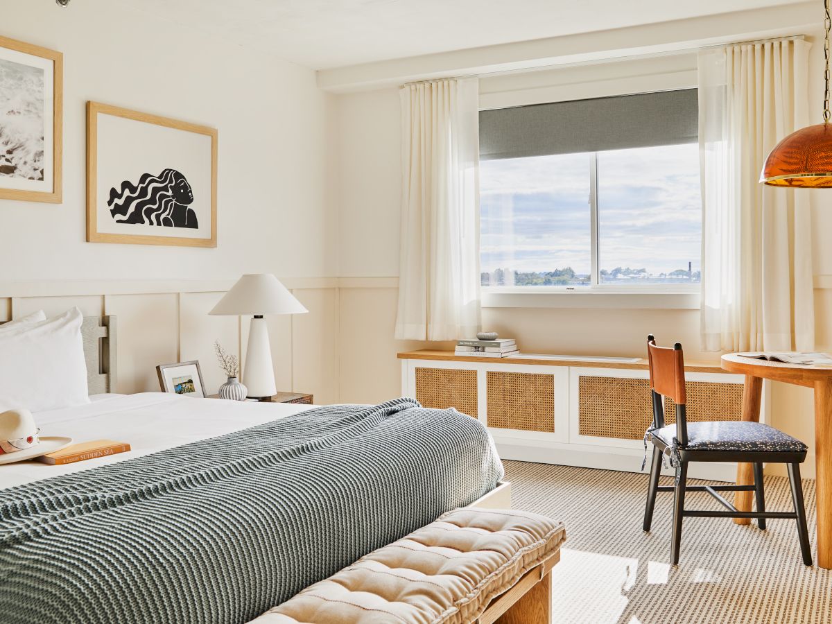 A cozy hotel room with a bed, artwork, a window, and a small dining area with a table and chair, all in a neutral color palette.
