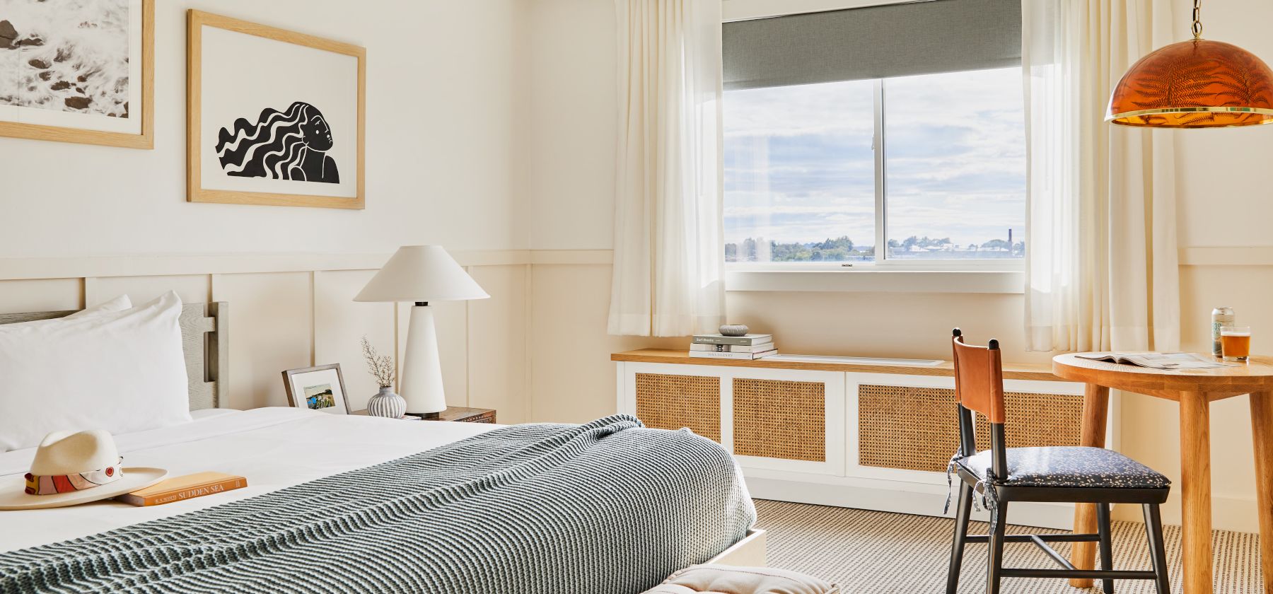 A cozy hotel room with a bed, artwork, a window, and a small dining area with a table and chair, all in a neutral color palette.