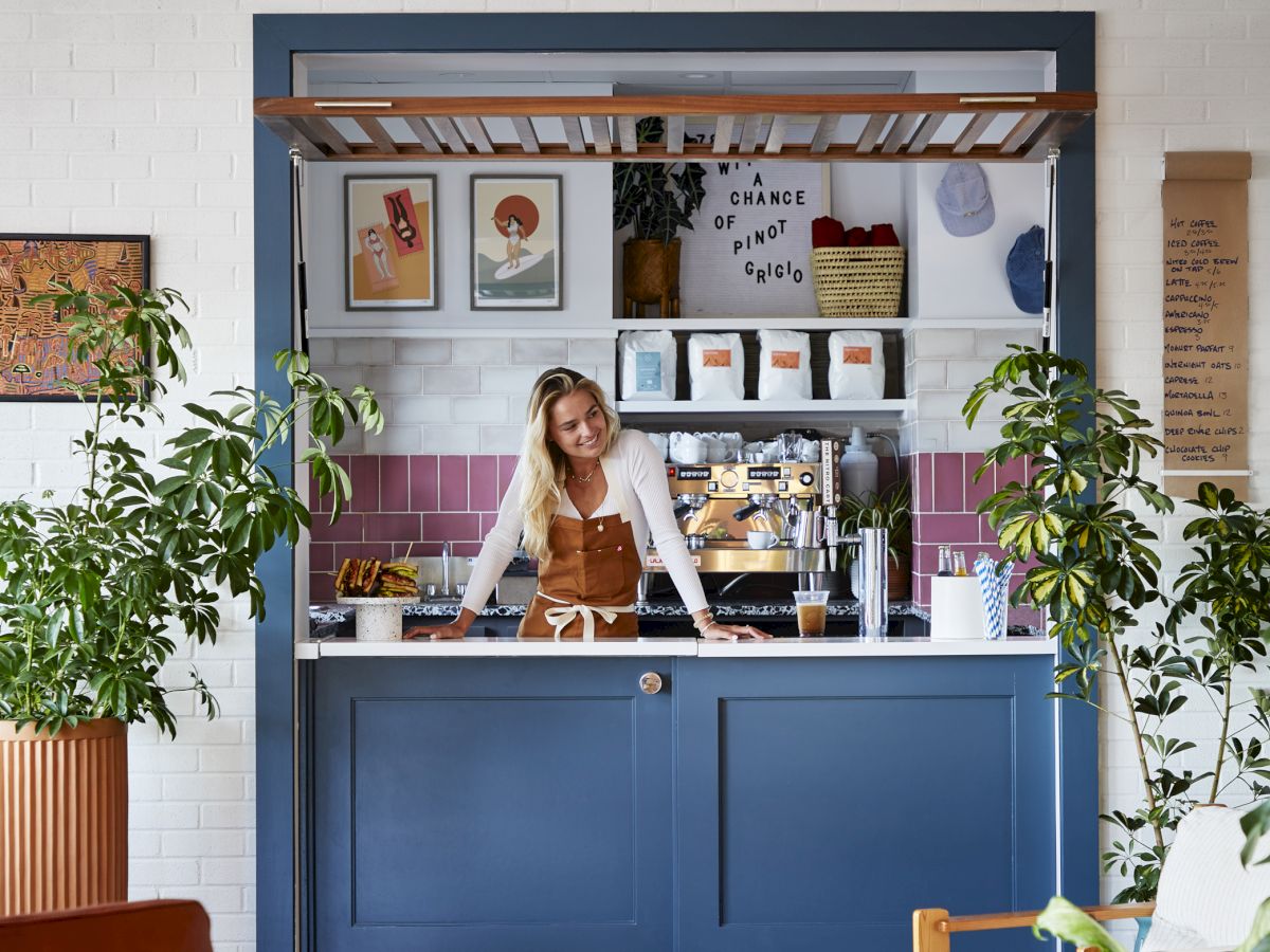 A cozy caf&eacute; corner with a woman standing behind a counter, surrounded by plants and colorful decorations, creating a welcoming atmosphere.