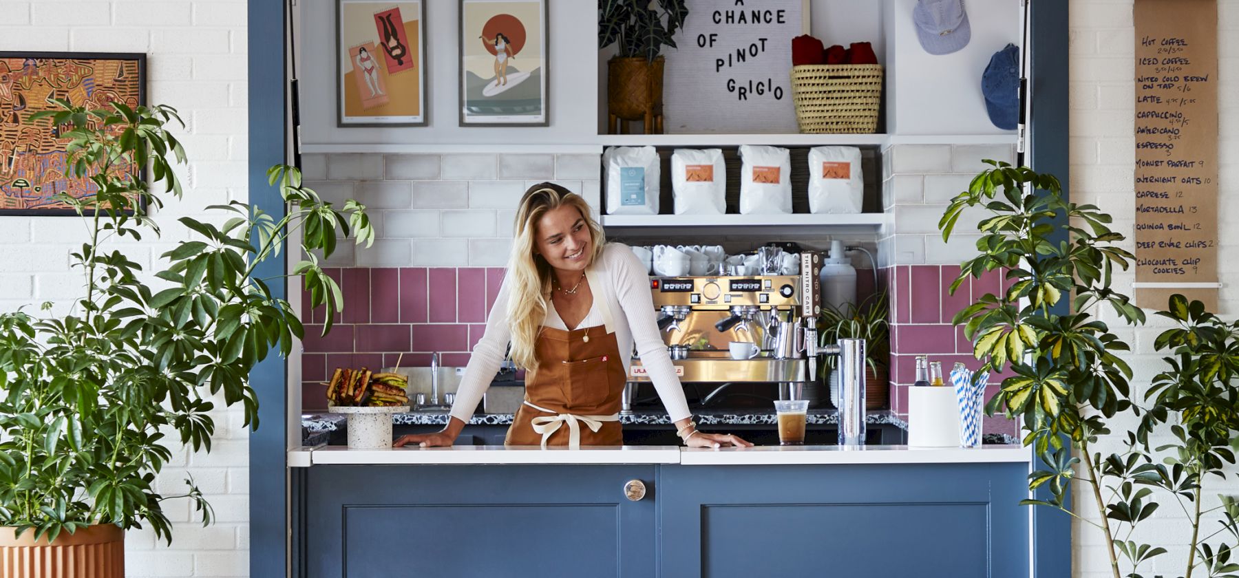 A cozy caf&eacute; corner with a woman standing behind a counter, surrounded by plants and colorful decorations, creating a welcoming atmosphere.