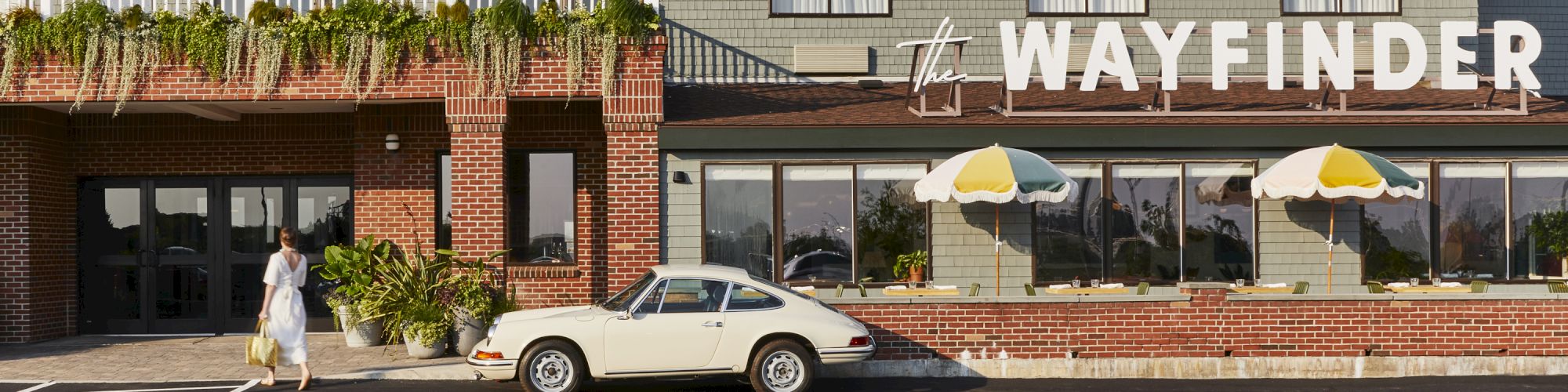 A person walks towards a vintage car parked outside a building labeled "The Wayfinder" with colorful umbrellas.